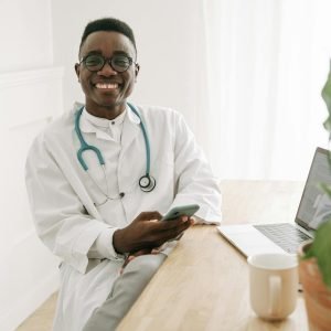 Happy doctor in uniform sitting at a desk using a smartphone in a bright office.