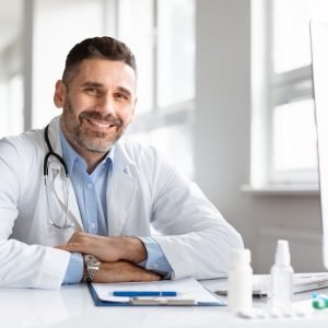Portrait of happy male doctor in white medical uniform, sitting at desk in hospital and smiling at